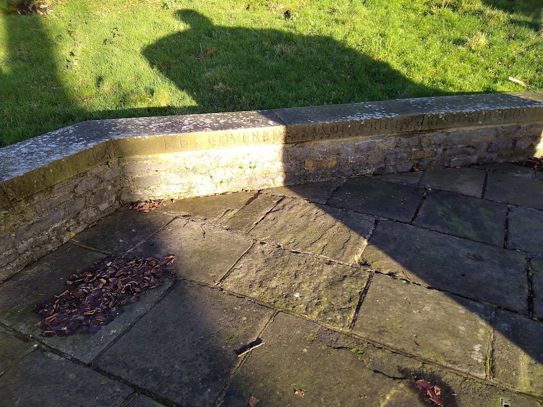 Stone seating area with a shadow cast on grass, surrounded by a paved path and scattered leaves.