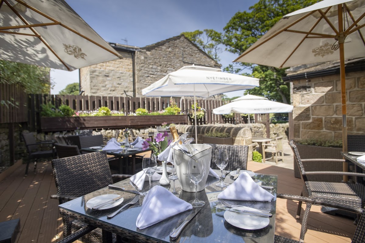 Outdoor dining area with a table set for guests, umbrellas, and stone buildings in a lush garden setting.
