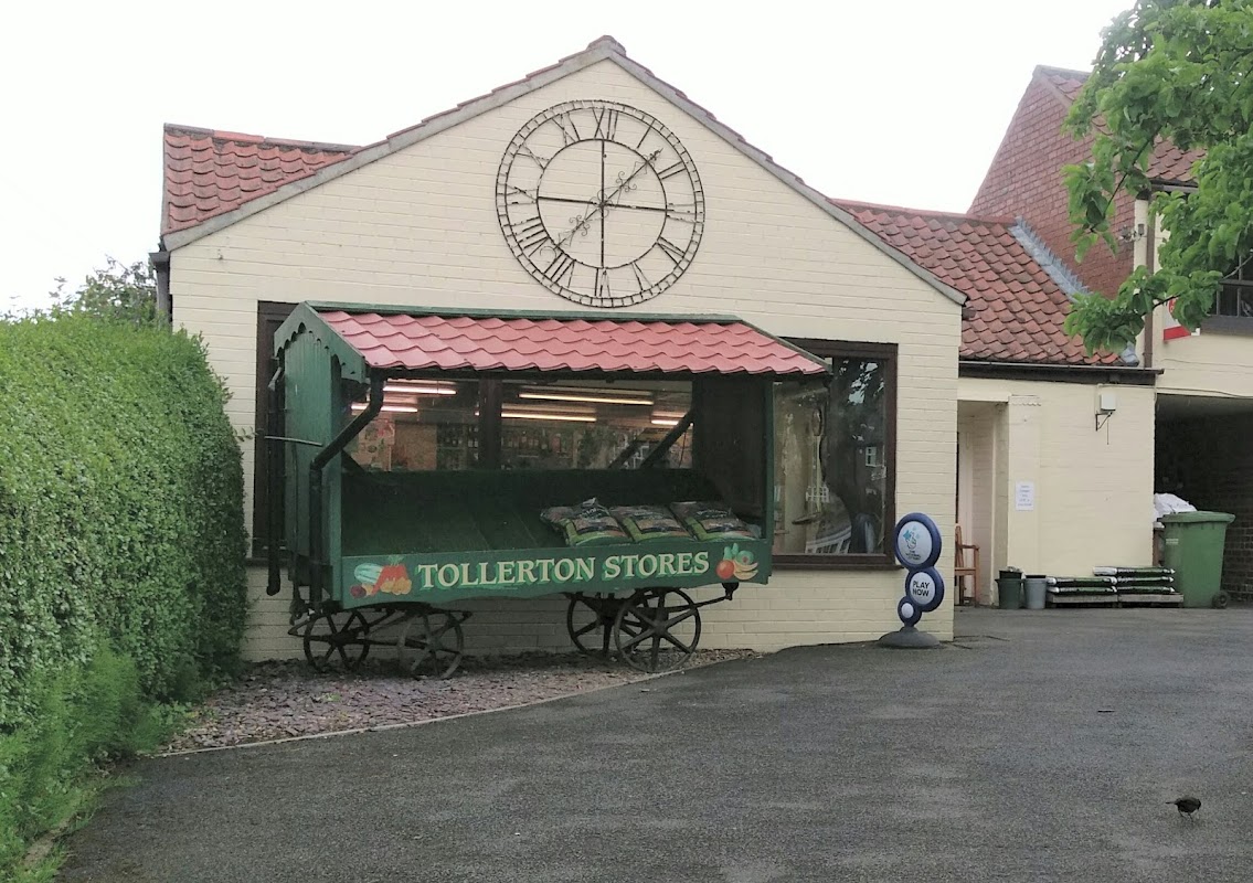 Tollerton Stores features a green cart display, large clock, and a brick building surrounded by greenery in Yorkshire.