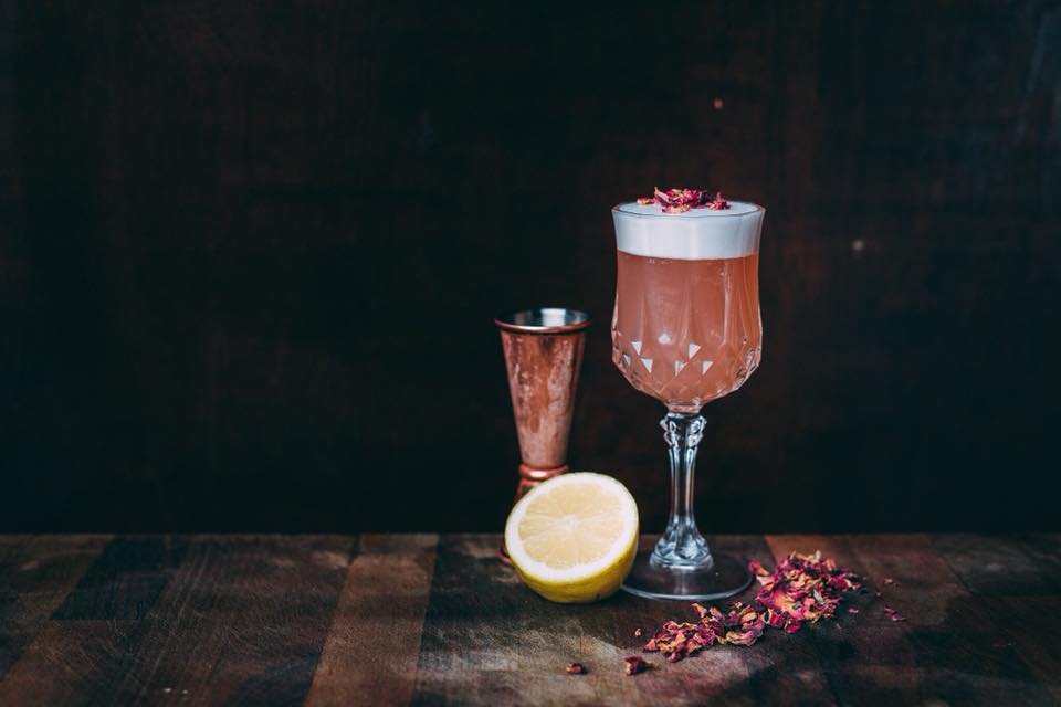 Elegant cocktail with foam, rose petals, and a lemon half on a wooden table at Vice and Virtue, Leeds.