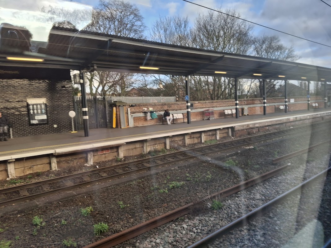 Wakefield Westgate Station platform with passengers, brick walls, and trees in the background under a cloudy sky.