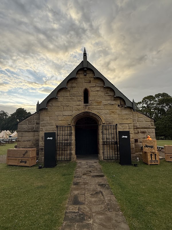 Historic stone building with a pointed roof, flanked by black columns and wooden crates on the lawn under a cloudy sky.