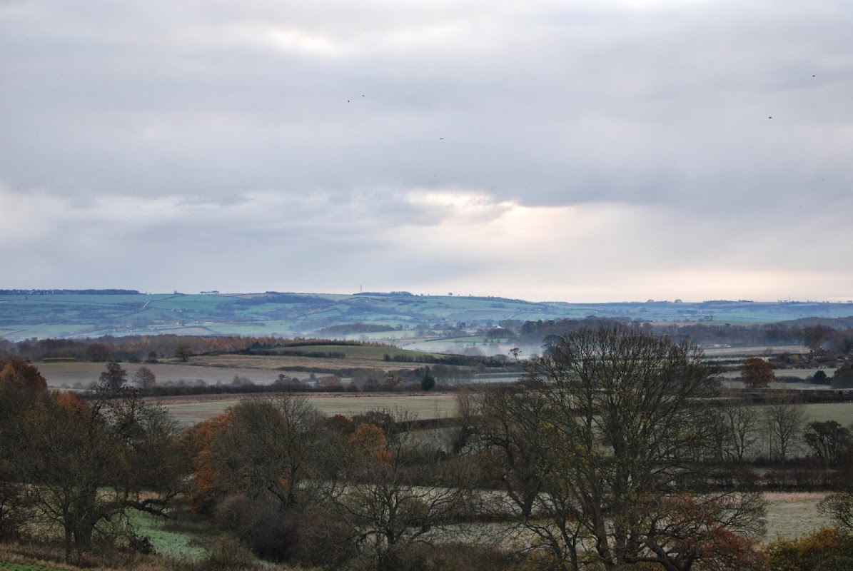 Welburn Village Hall surrounded by rolling hills and trees under a cloudy sky in the Yorkshire countryside.