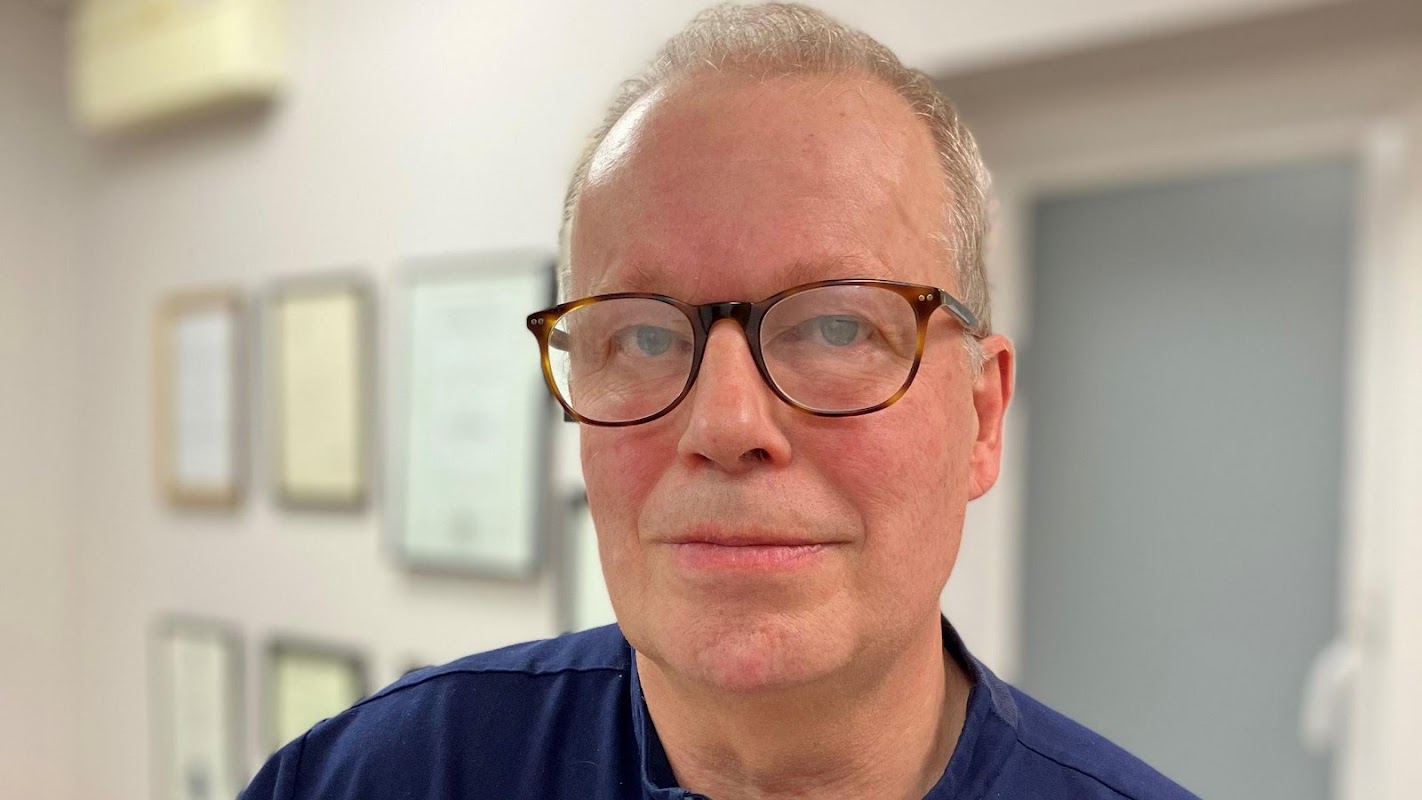 Dr. Paul Charlson wearing glasses, in a clinic setting with framed certificates in the background.