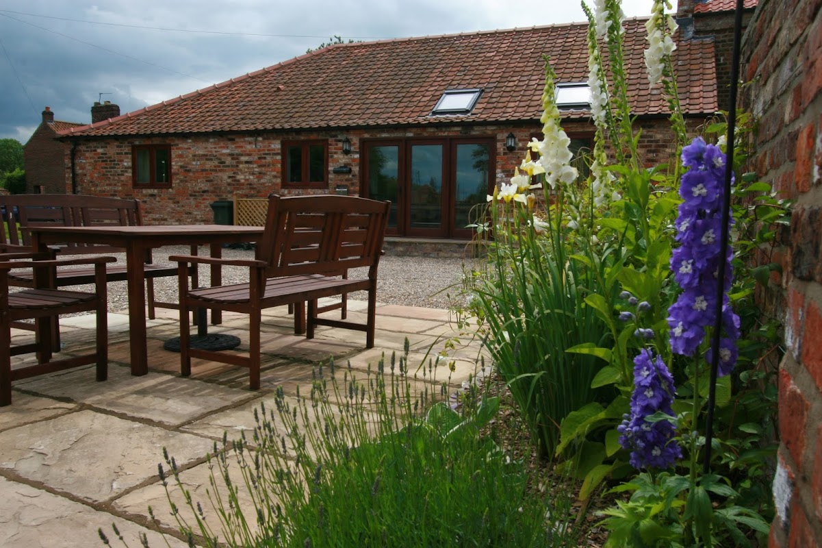 Rustic holiday cottage with a stone patio, wooden seating, and vibrant flowers in West Lutton, Yorkshire.