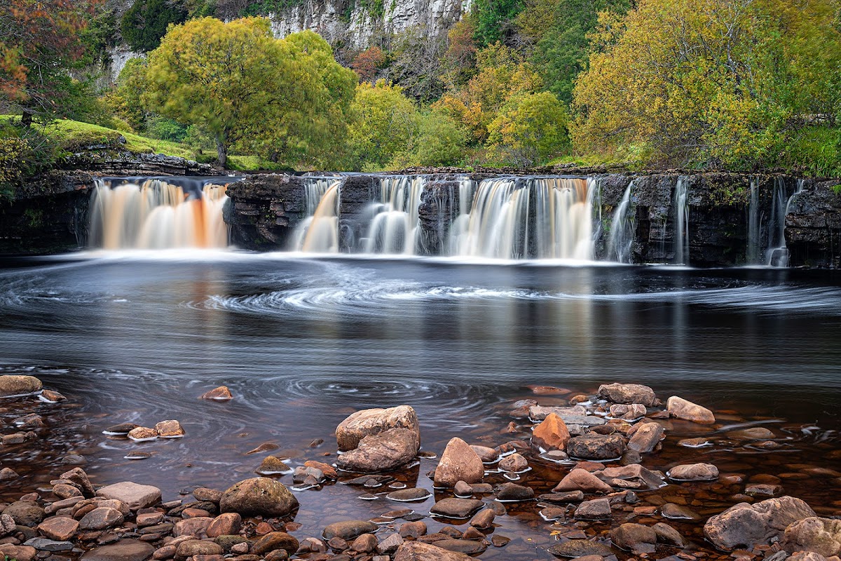 Waterfall cascading over rocks, surrounded by vibrant autumn foliage and a serene river in West Stonesdale, Yorkshire.