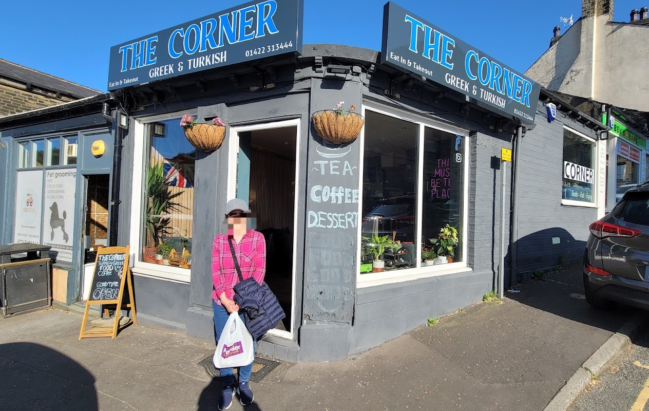 A woman stands outside "The Corner," a Greek and Turkish restaurant with a blue sign and potted plants.