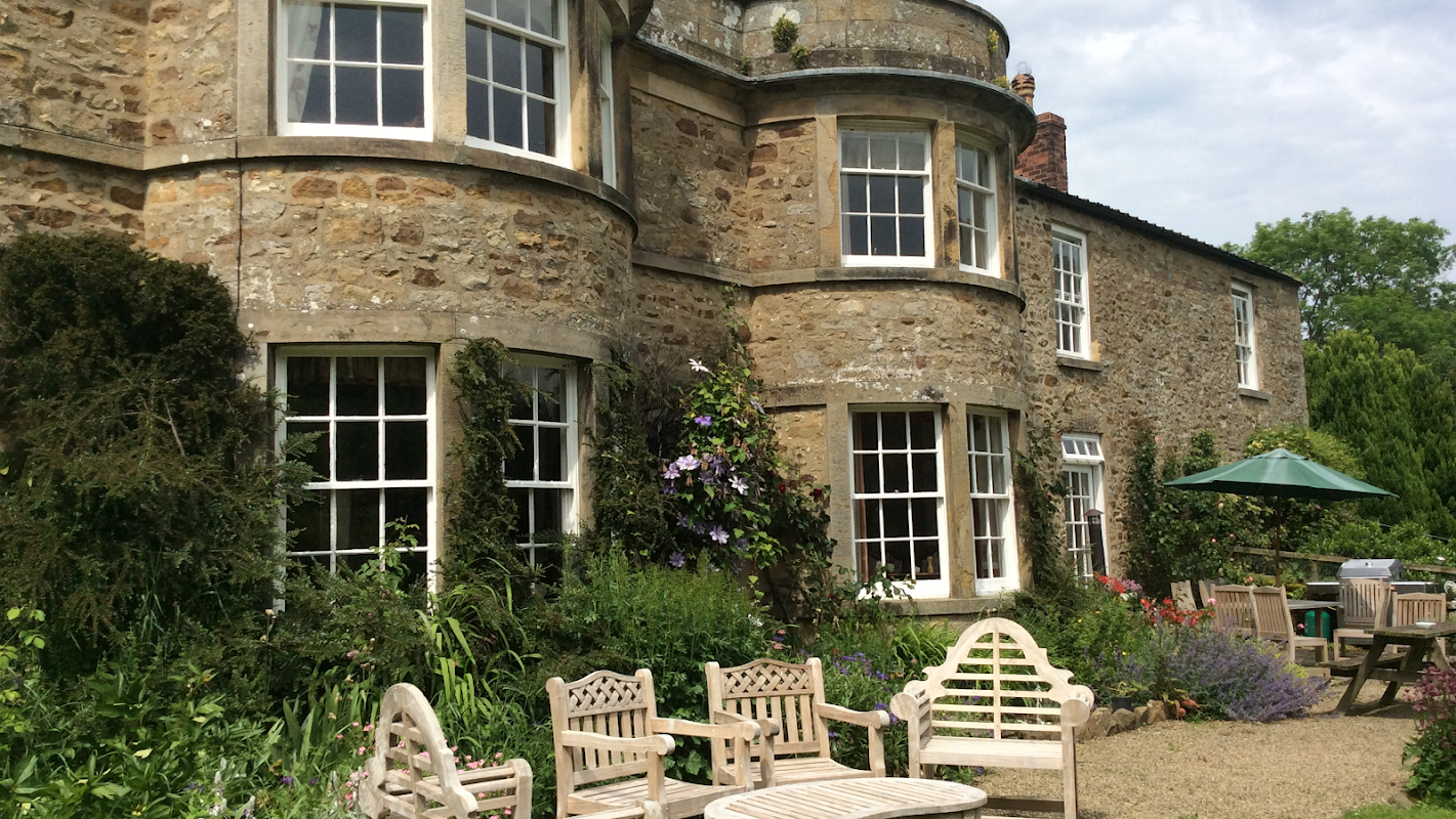 Stone farmhouse with large windows, surrounded by lush greenery and outdoor seating in a tranquil garden setting.