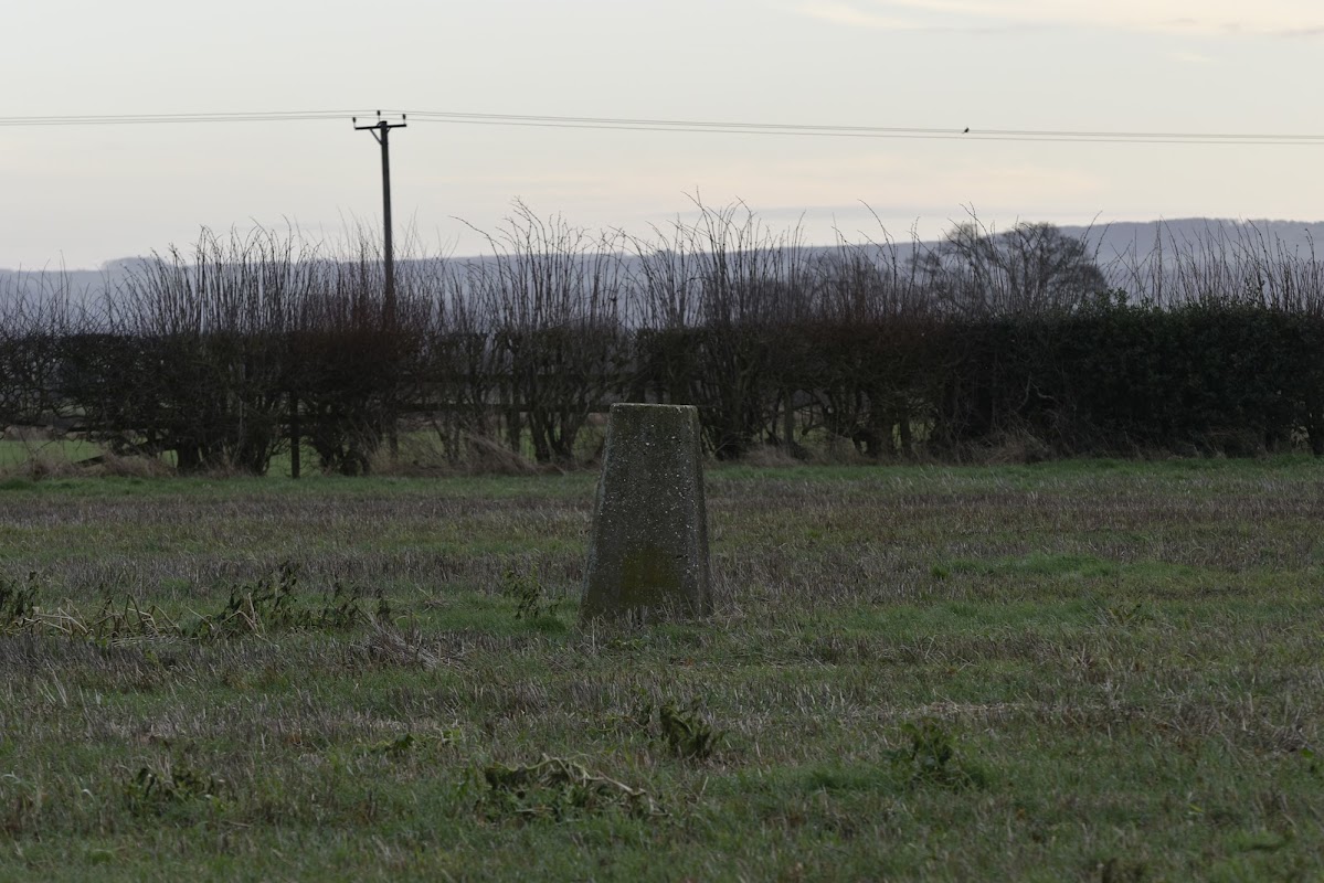 Trig point on a grassy field, surrounded by hedgerows and distant hills under a soft sky.