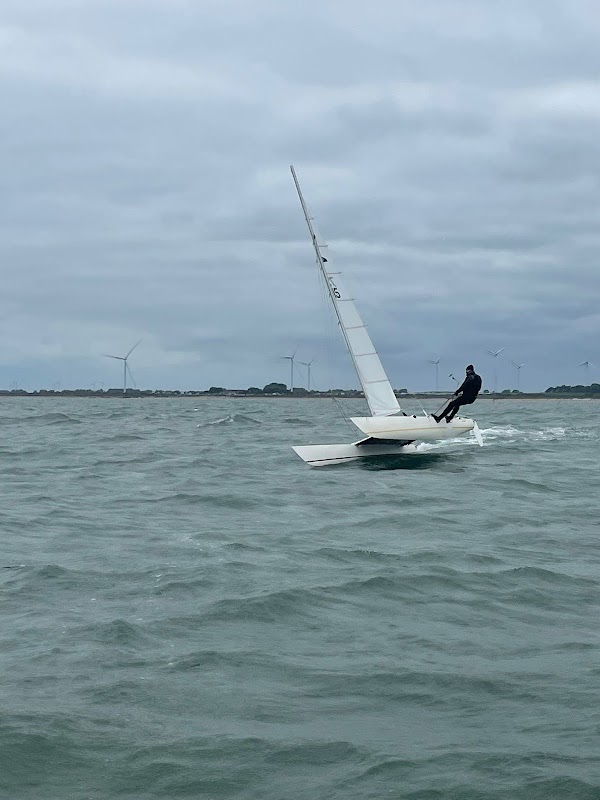 A sailor on a small dinghy navigating choppy waters, with wind turbines visible in the background under a cloudy sky.