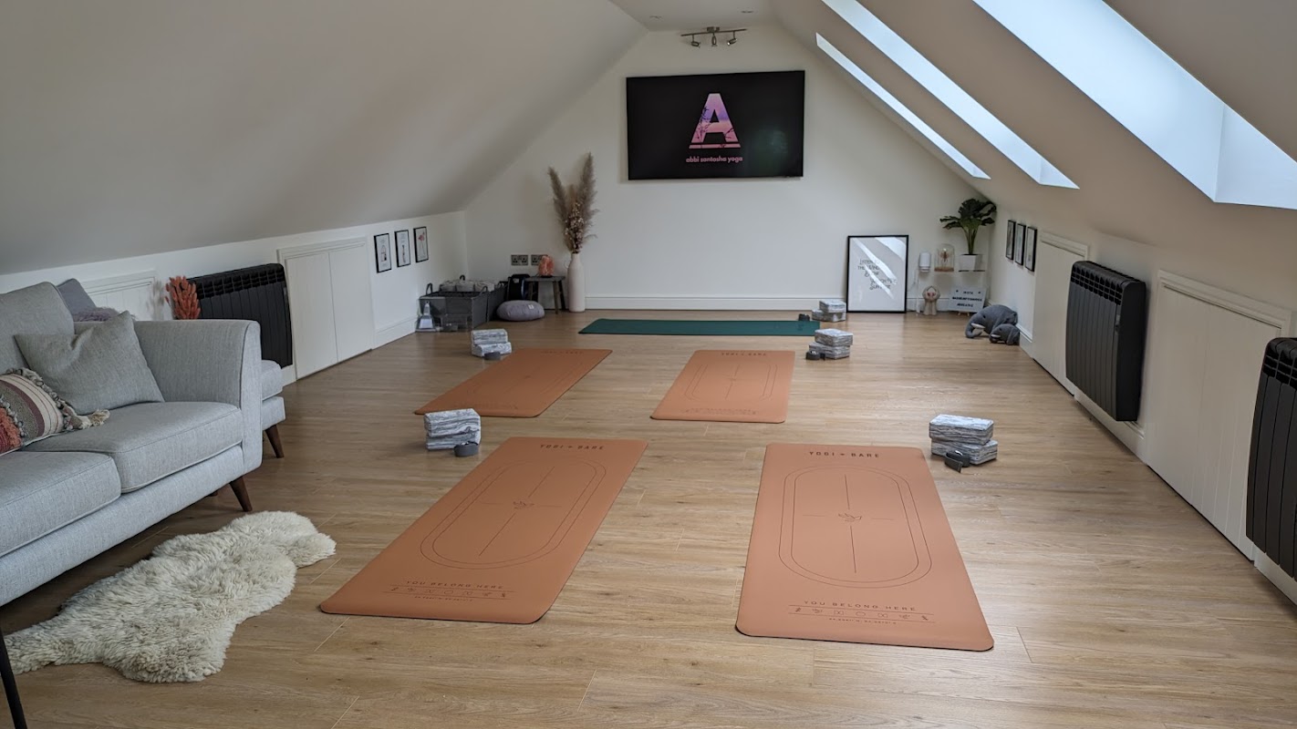 Yoga studio with mats arranged on wooden floor, cozy seating area, and a screen displaying "Abbi Santosha Yoga."
