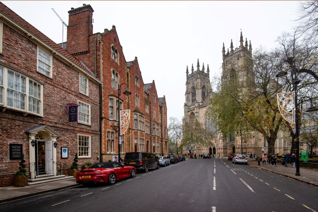 Historic buildings line a street, with Dean Court Hotel and York Minster's towering spires in the background.