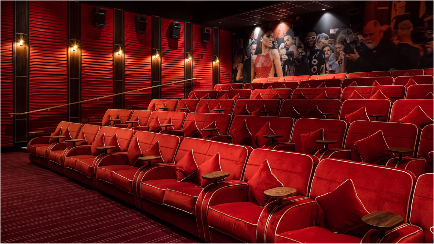 Red velvet seating with armrests and cushions, arranged in rows, illuminated by soft wall lights in a theater setting.