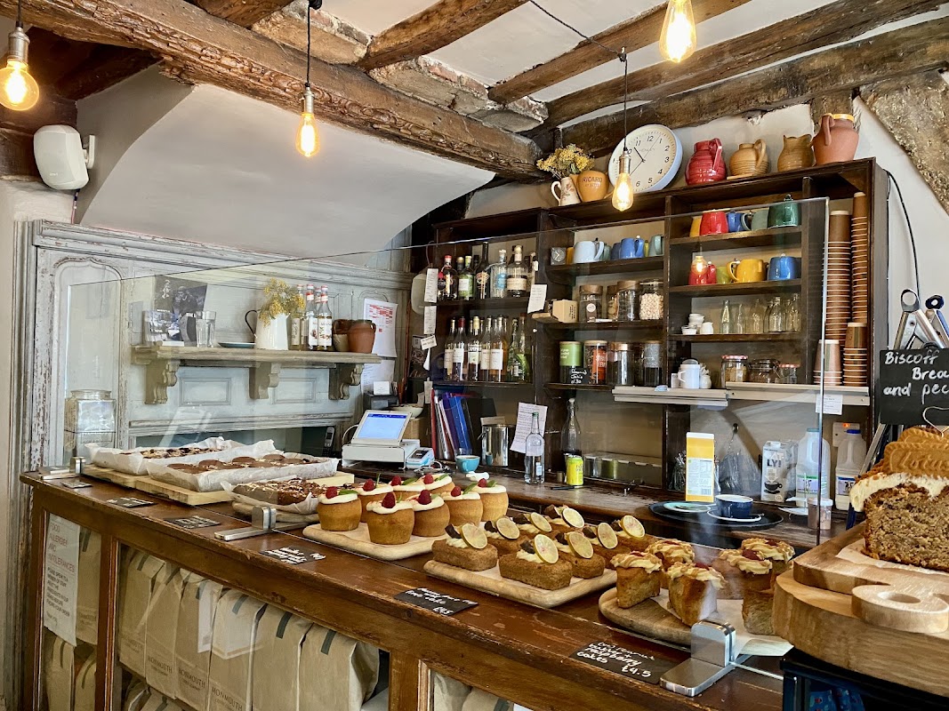 Cozy cafe interior with a wooden counter, baked goods, colorful mugs, and a rustic shelf filled with jars and bottles.