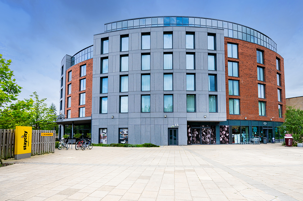 Modern hotel building with a mix of grey and red brick, large windows, and a spacious plaza with bicycles parked outside.