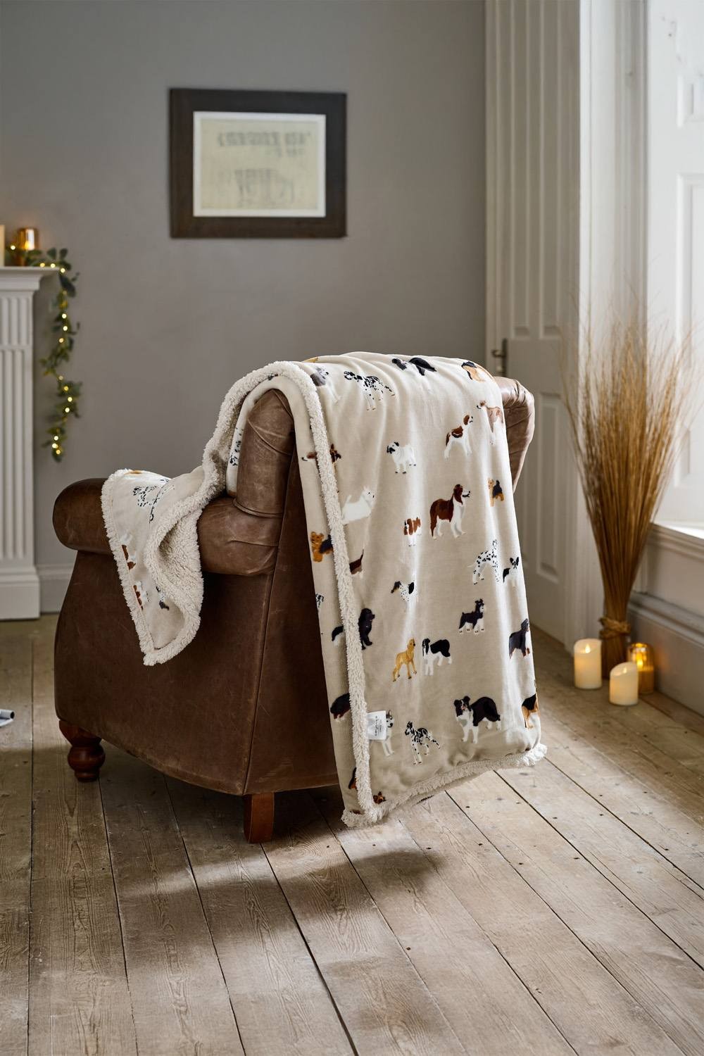 Beige fleece throw featuring mixed dog patterns, soft Sherpa backing, measuring 140x180cm, draped over a brown leather armchair in a cozy interior setting.