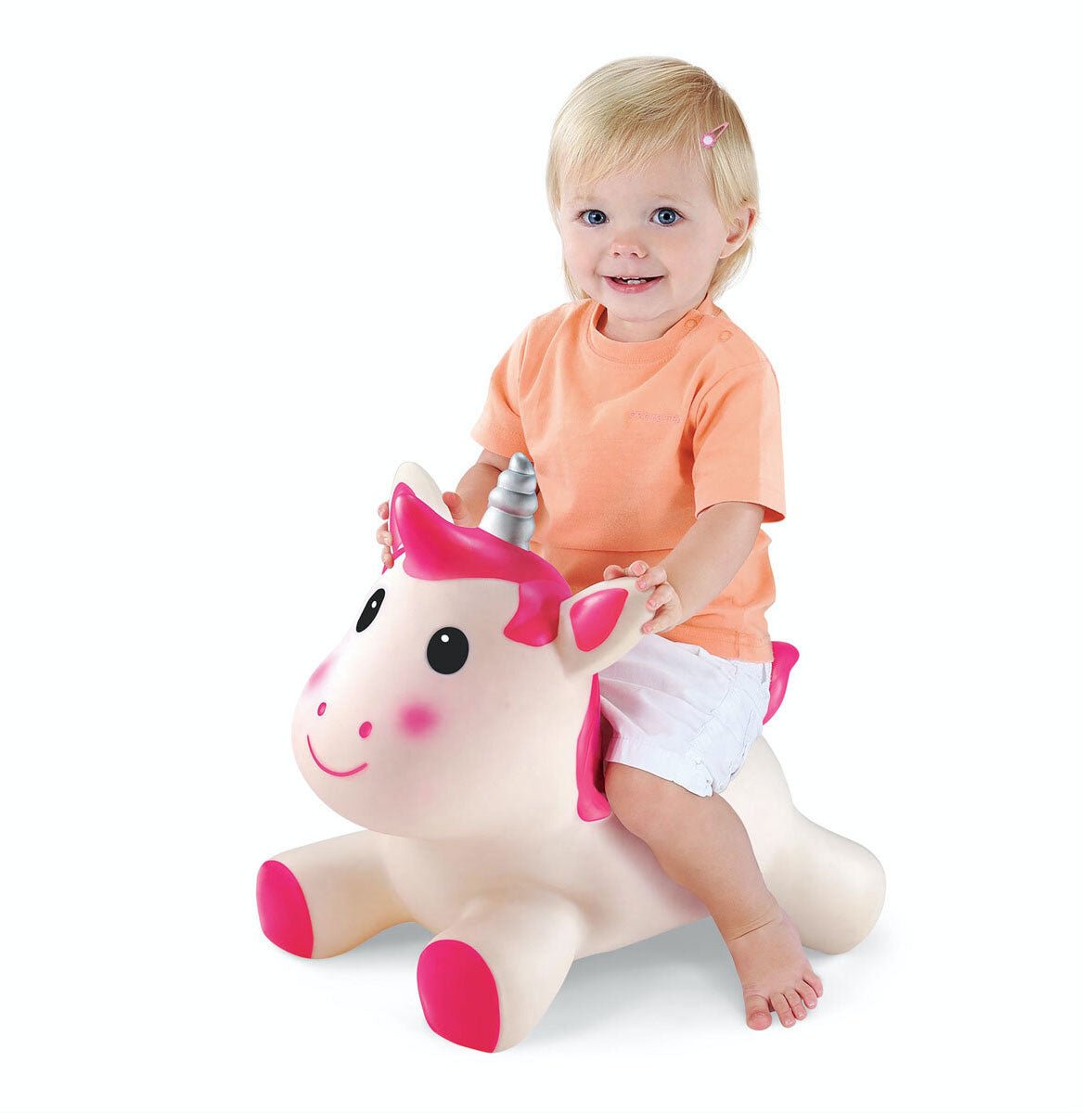 A young child with blonde hair sits on a soft, pink and white unicorn toy with a smiling face and a silver horn, wearing an orange shirt and white shorts.