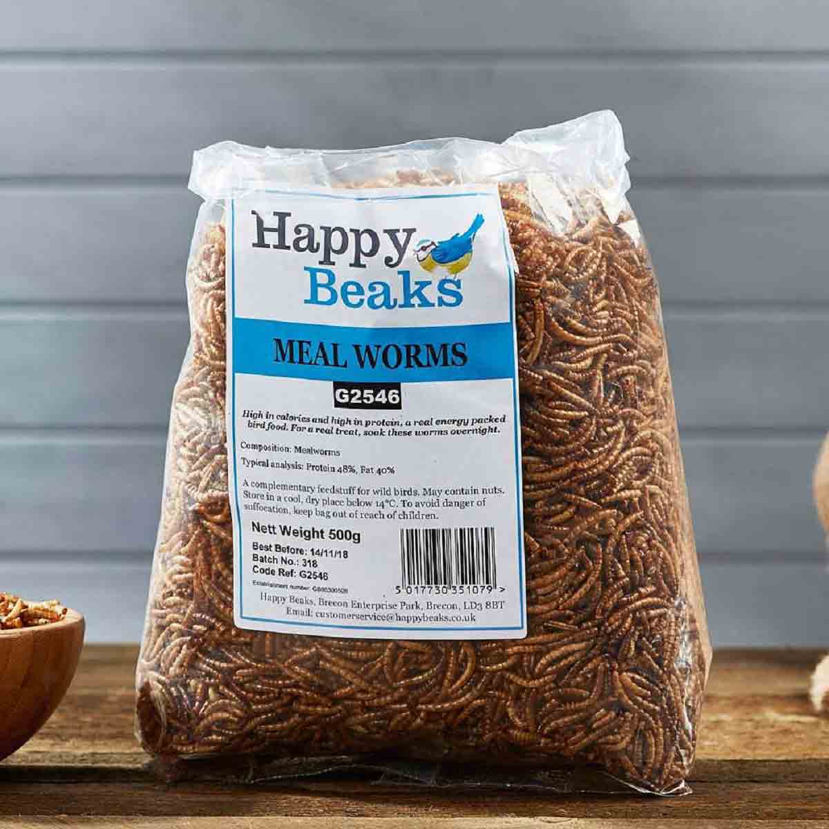 Clear plastic bag containing 1 kg of Happy Beaks meal worms, featuring a blue and white label with product details, set against a wooden surface.