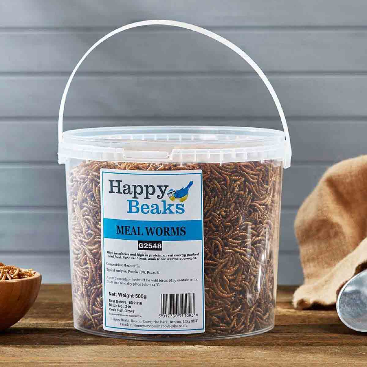 Clear plastic tub containing 1 kg of meal worms, labeled "Happy Beaks" in blue and white, with a handle and a brown cloth in the background.