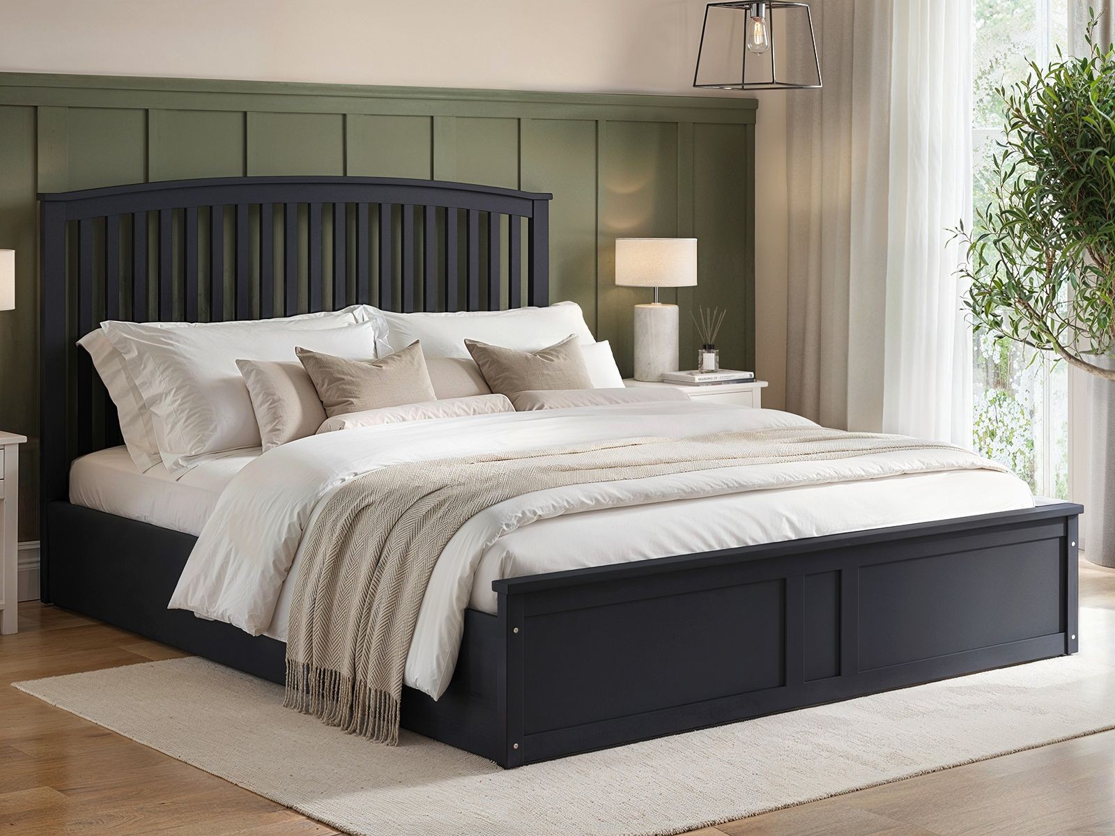 Wharfedale Wooden Ottoman Bed by Flair features a dark navy frame, slatted headboard, and a spacious storage base, complemented by soft bedding and neutral decor in a bright room.
