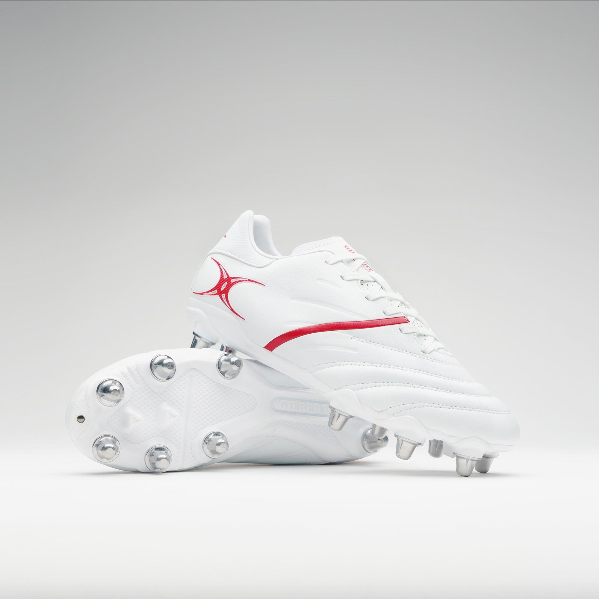 White rugby boots with red accents, featuring a sleek design, textured upper, and metal studs on the sole for traction, designed for senior players by Gilbert Rugby.