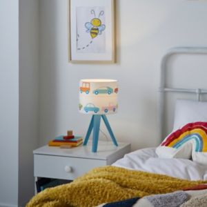 White lampshade with colorful car illustrations on a blue wooden tripod base, placed on a bedside table next to a cozy blanket and a rainbow pillow.