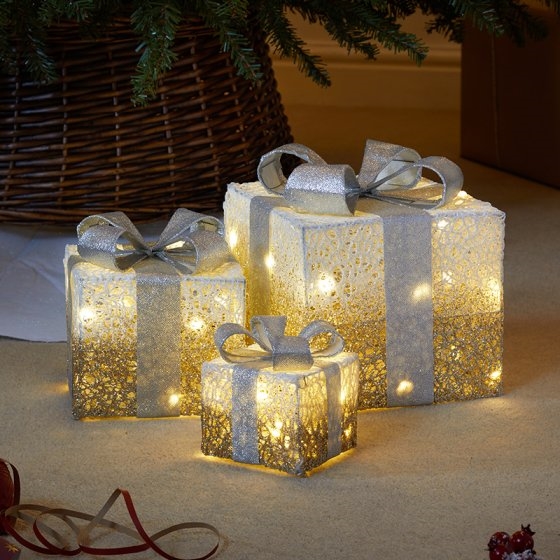 Three sparkly faux gift boxes in gold and silver, adorned with ribbons and lights, varying in size, set against a warm, festive backdrop.