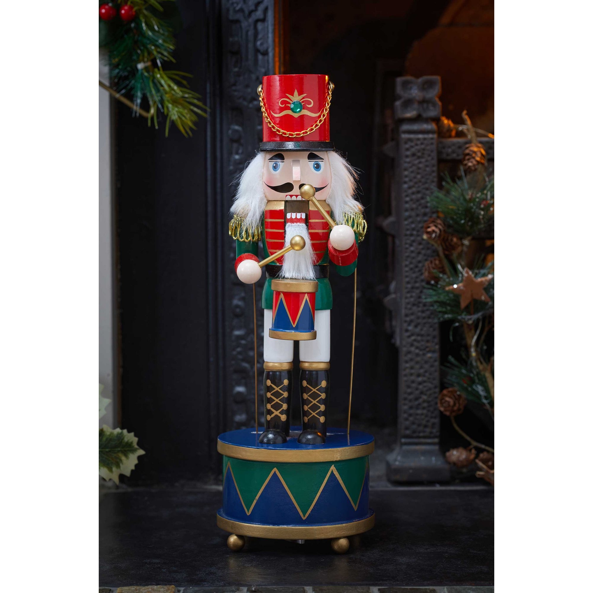 Colorful wooden nutcracker with a red hat, white beard, and green and blue uniform, holding drumsticks, standing on a round base with geometric patterns.