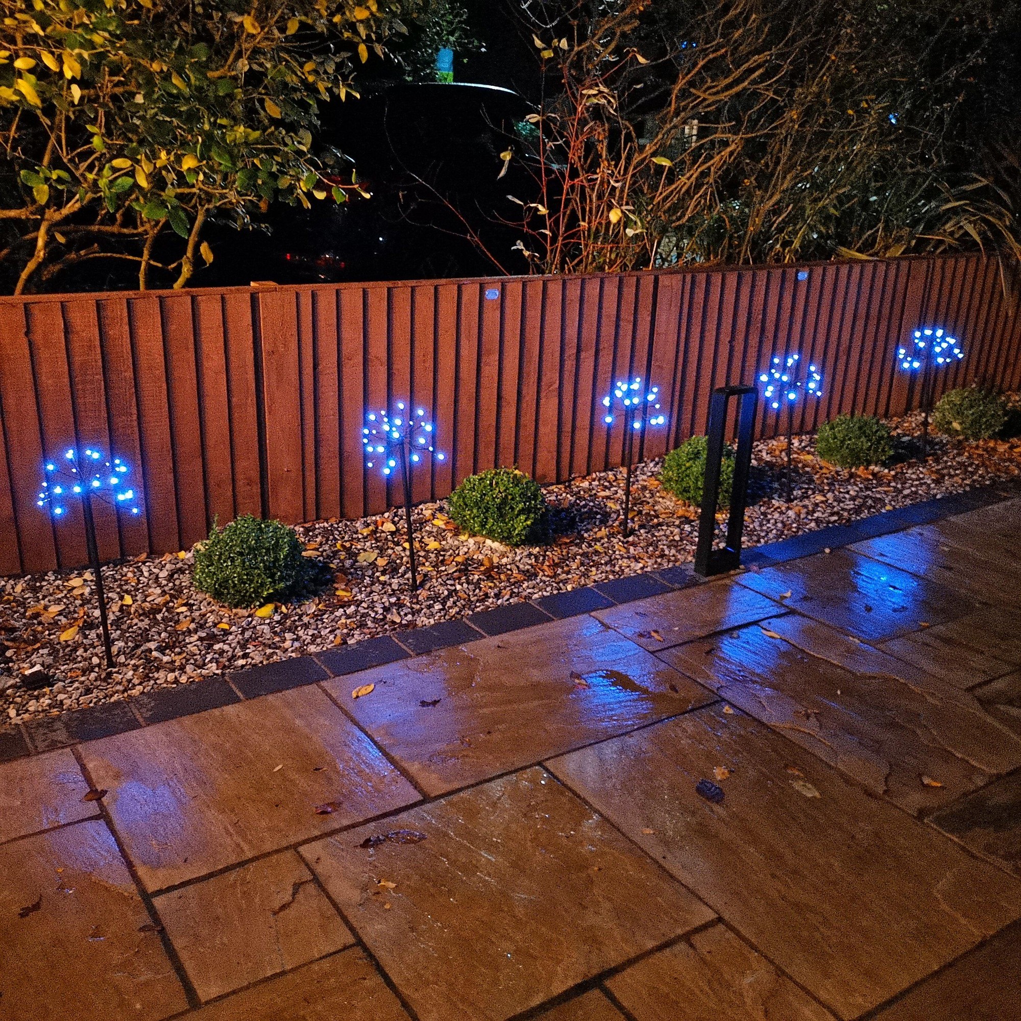 Five battery-operated sparkler pathfinder lights with 120 cool white LEDs, featuring a sleek design, placed along a stone pathway next to green shrubs and wooden fencing.