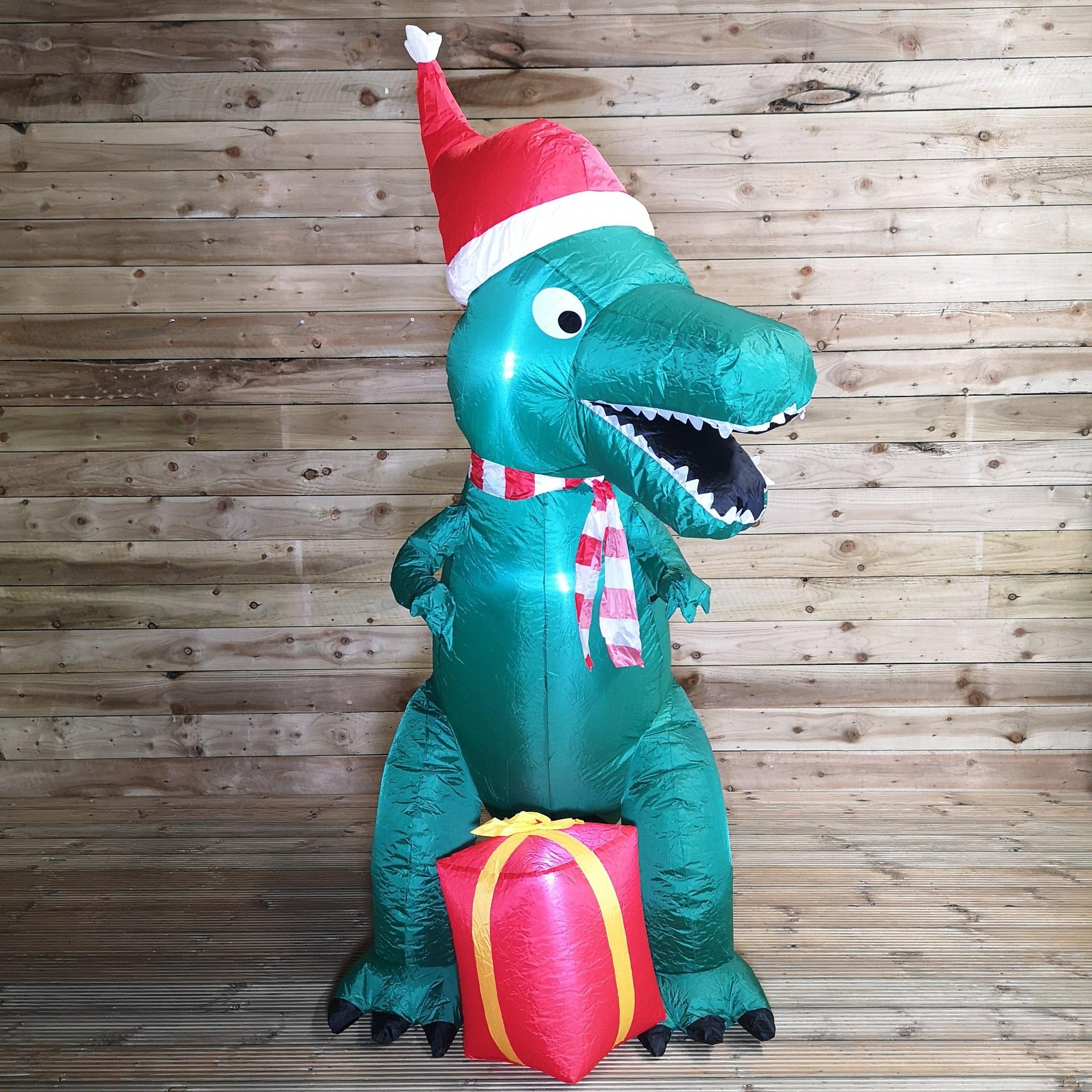 Green inflatable T-Rex dinosaur wearing a red Santa hat and a striped scarf, sitting beside a red parcel, against a wooden background.