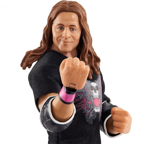 Bret "Hit Man" Hart wrestling action figure features long brown hair, a black shirt with a skull design, and a pink wristband, posed with a clenched fist.