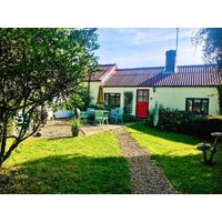 Charming yellow cottage with red door, patio furniture, and lush garden, surrounded by trees and a gravel path.