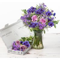Bouquet of purple and pink flowers in a glass vase, with matching blooms in an open Appleyard box nearby.