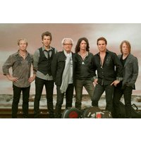 Band Foreigner standing together, six members in casual and formal attire, with a cloudy sky backdrop.