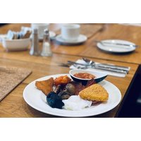 Plate of English breakfast with hash brown, sausage, bacon, and poached egg on wooden table with cutlery and cups.