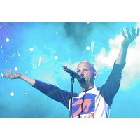 Singer on stage with microphone, wearing a blue and white shirt, arms raised against a backdrop of blue lights and smoke.