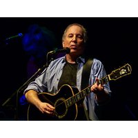 Musician playing black acoustic guitar, wearing a blue shirt over a black t-shirt, singing into a microphone on stage.