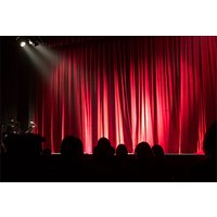 Red theater curtains illuminated by spotlight, with silhouetted audience in foreground.