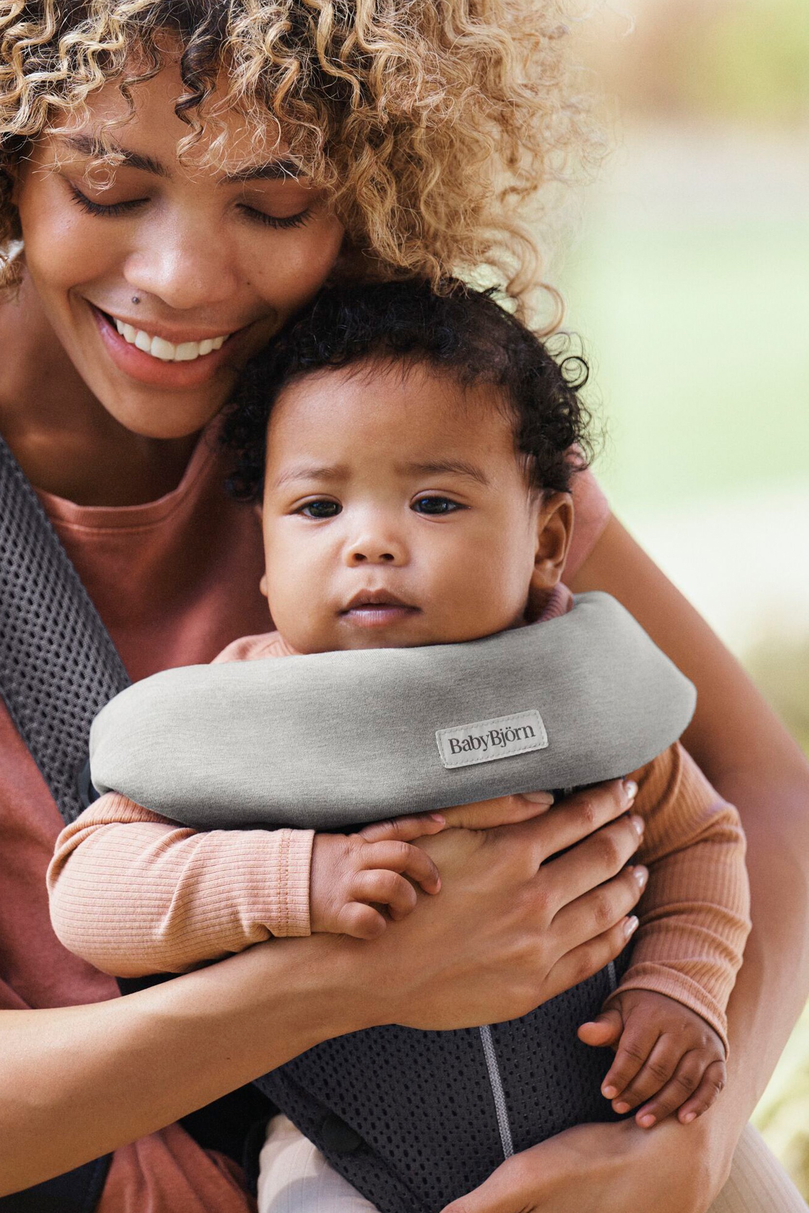 Light grey jersey bib designed for BabyBjorn Mini and Move carriers, featuring soft fabric and a logo patch, worn by a parent holding a baby.