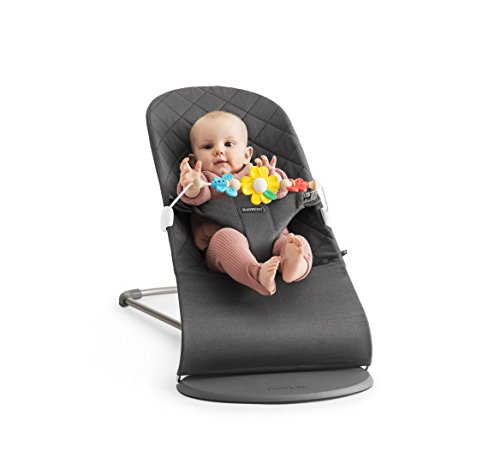 A baby bouncer in dark gray fabric with a quilted pattern, featuring a colorful flower toy bar, and a seated baby wearing a pink outfit with legs visible.