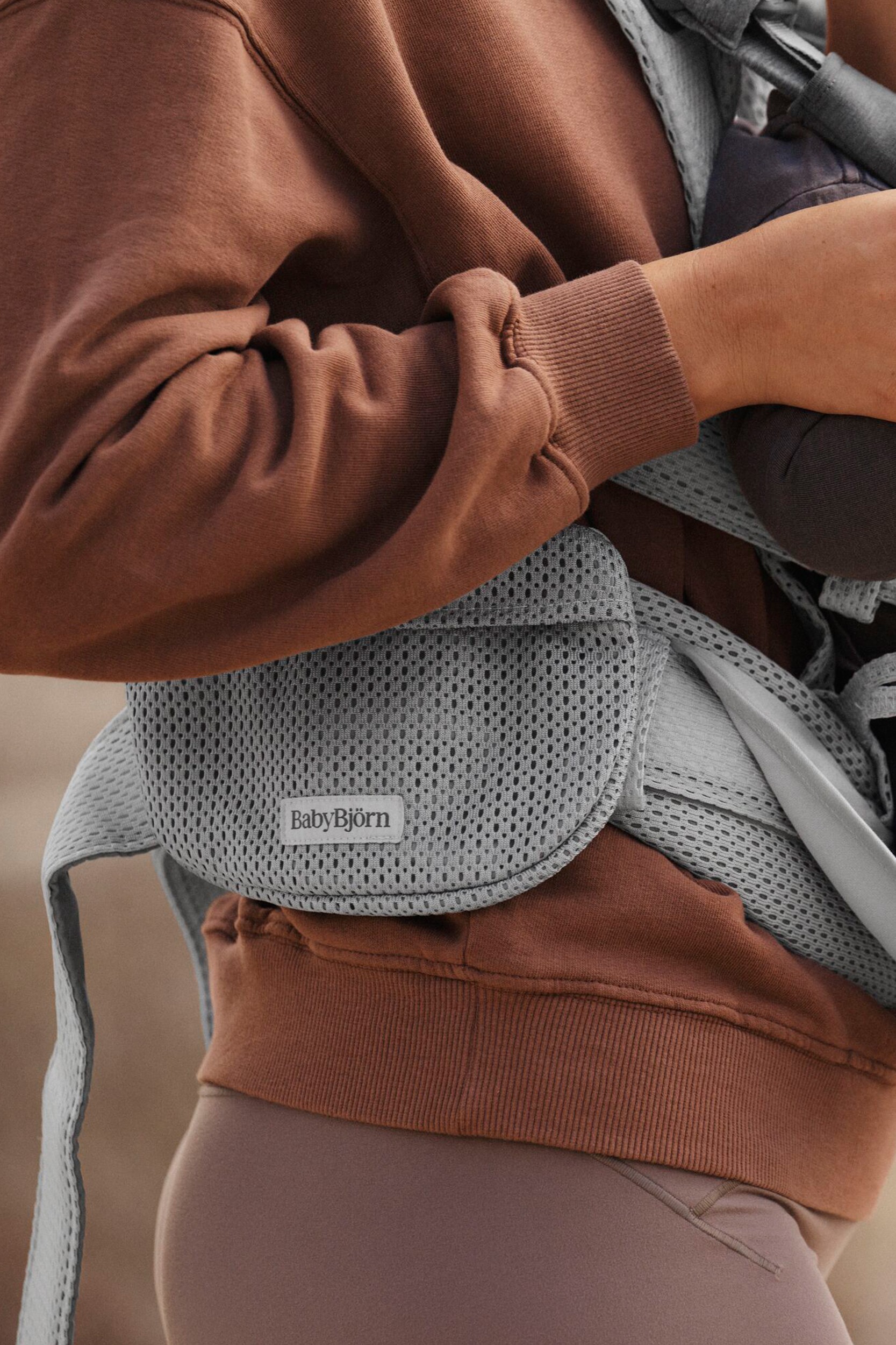 Grey mesh pocket pouch for BabyBjorn baby carrier, featuring adjustable straps and a logo tag, attached to a brown sweatshirt.