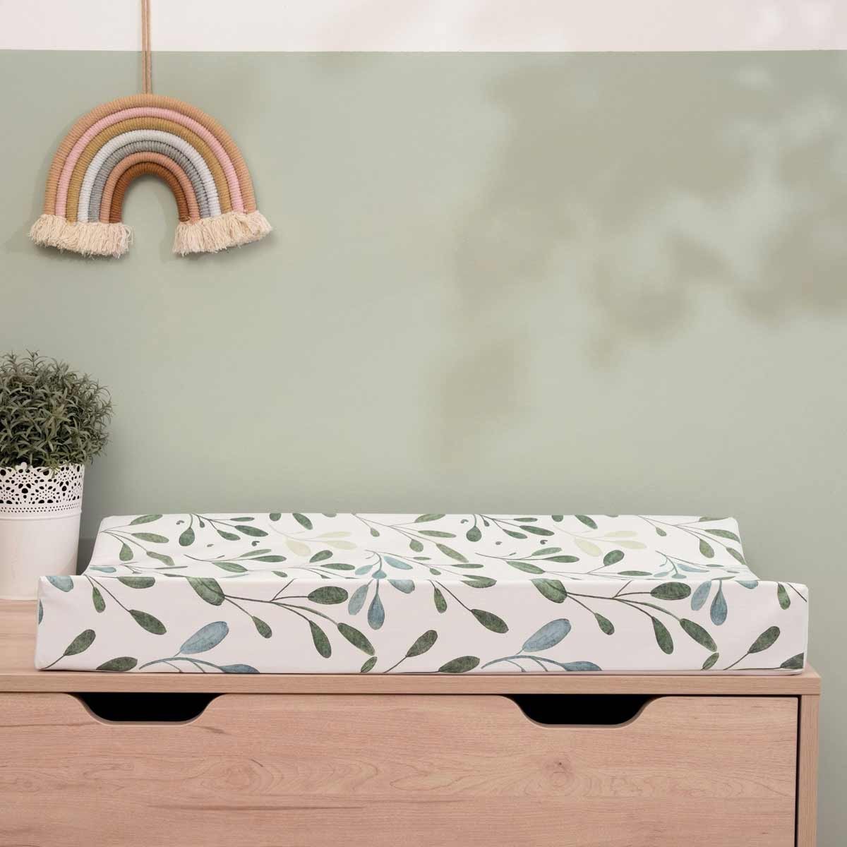 Olive-patterned changing mat in green and white with anti-roll wedge design, placed on a wooden dresser, accompanied by a potted plant and a rainbow wall decor.