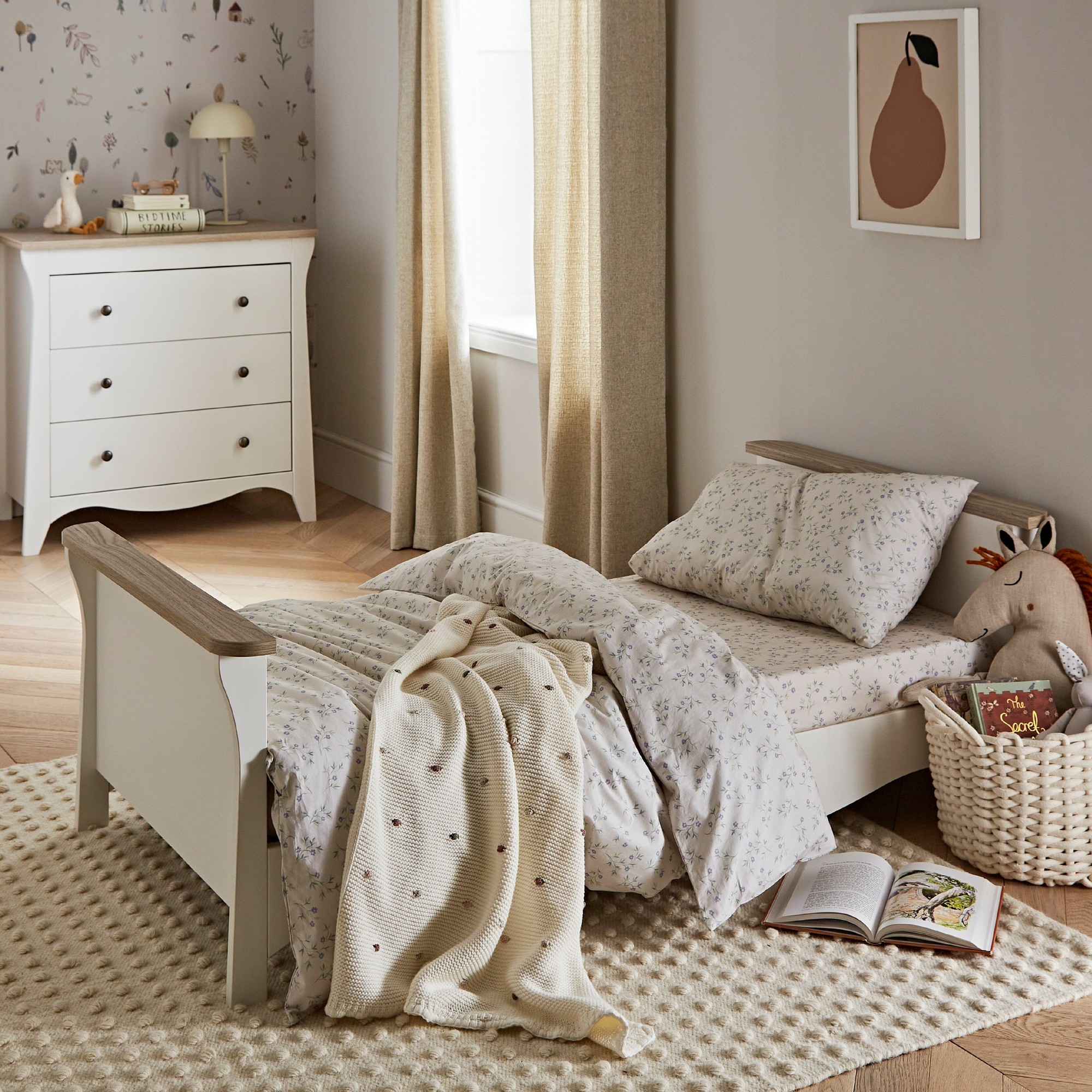 Clara Toddler Bed in white and ash features a low profile, wooden frame with a soft, neutral aesthetic, complemented by floral bedding and a cozy blanket on a textured rug.