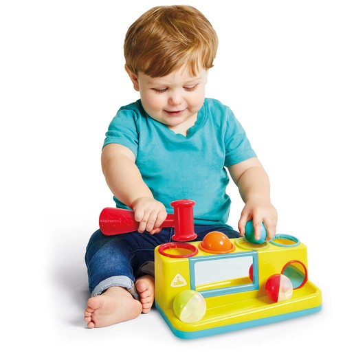 A young child in a teal shirt plays with a bright yellow hammer ball bench toy, featuring colorful shapes and a red hammer for striking the balls.