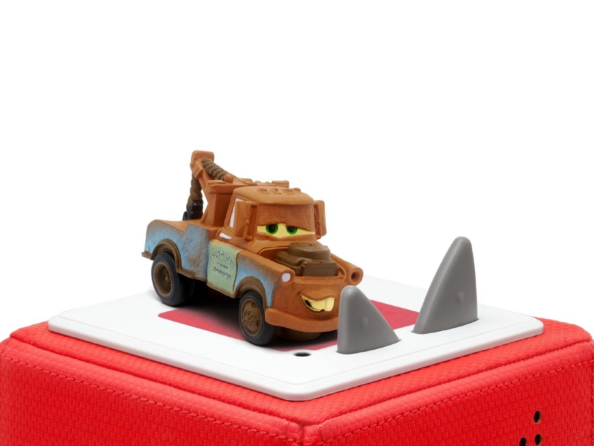 A brown and blue toy tow truck with a smiling face and green eyes sits on a red cube speaker, featuring two gray cones at the front.