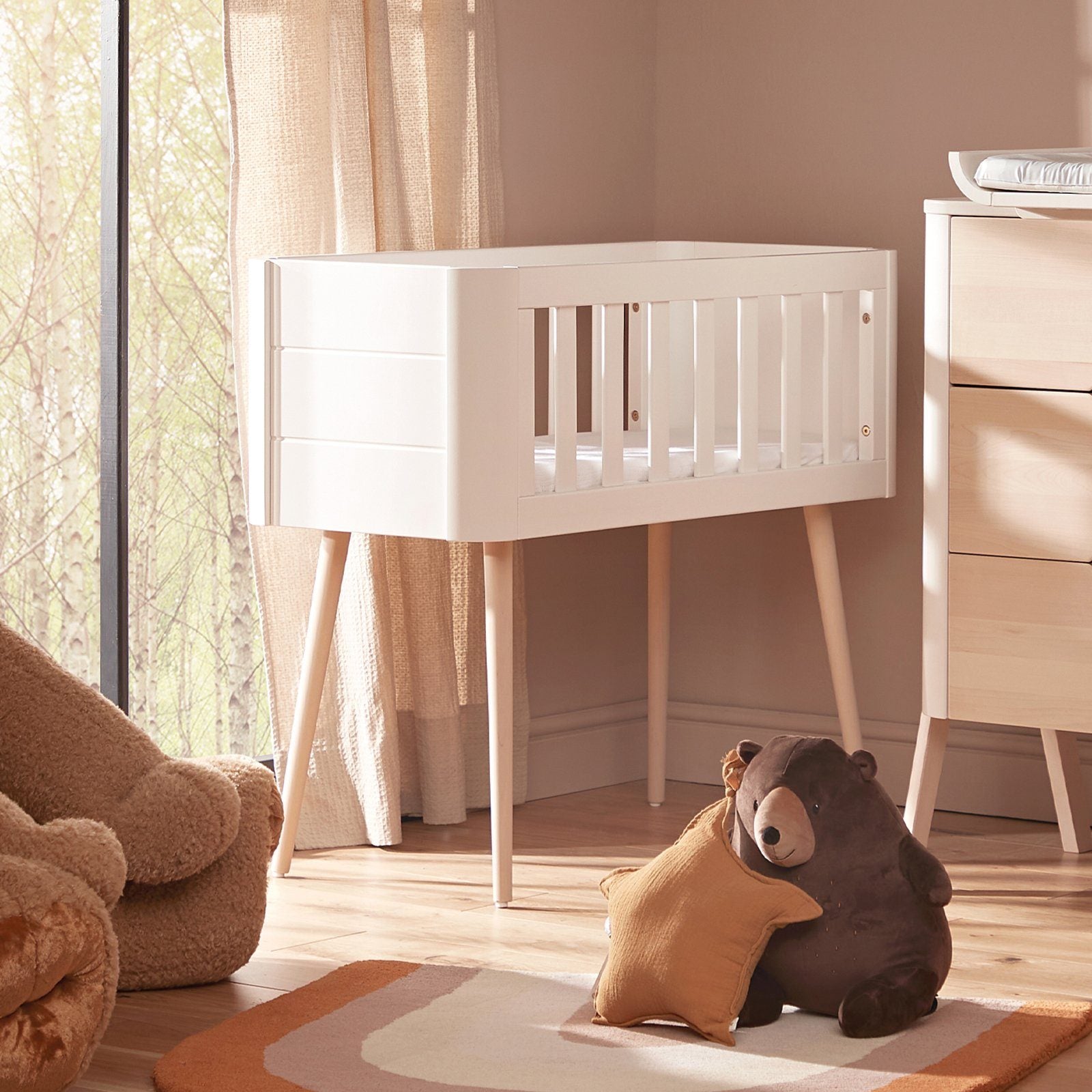 White and natural wood retro crib with slatted sides, standing on tapered legs, beside a plush bear and a colorful rug in a softly lit nursery.