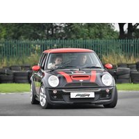 Black Mini Cooper with red roof and racing stripes on a track, two people inside, surrounded by tires and greenery.