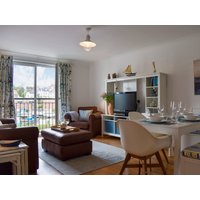Modern living room with brown leather sofa, white dining table, TV on white shelf, and nautical decor by large window.