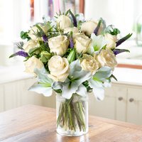 Bouquet of cream roses, purple veronica, and eucalyptus in a clear glass vase on a wooden table.