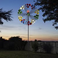 Colorful garden wind spinner with red, green, and yellow petals on a tall metal pole against a sunset backdrop.